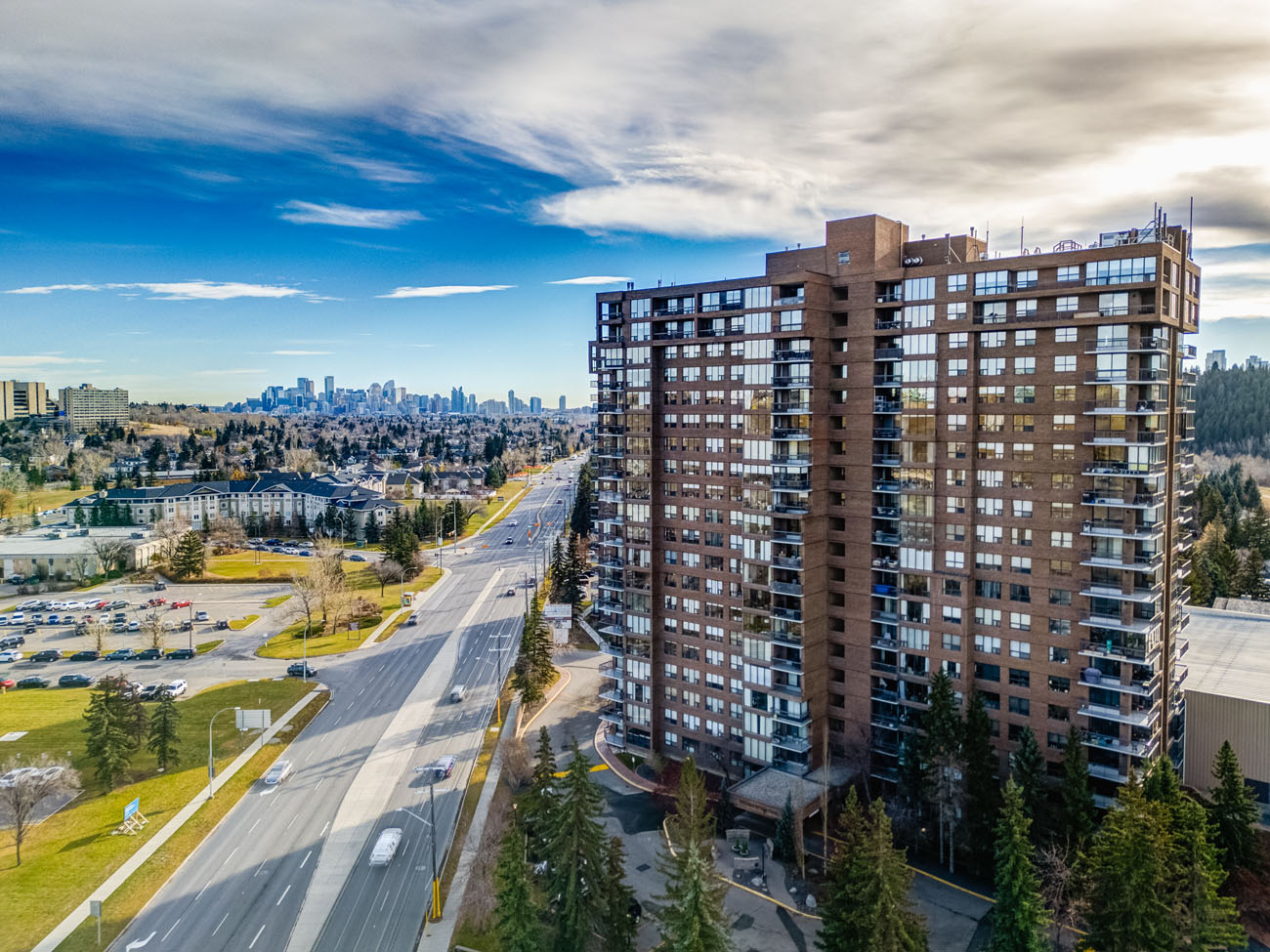 Aerial drone photograph looking down a Calgary boulevard toward a high-rise residential tower and the downtown skyline